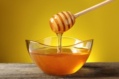 Delicious honey flowing down from dipper into bowl on wooden table against yellow background
