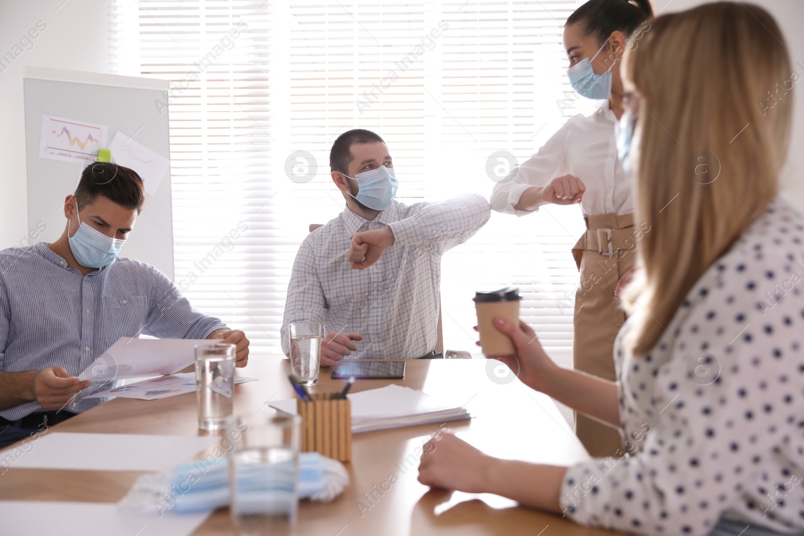 Photo of Coworkers with protective masks making elbow bump in office. Informal greeting during COVID-19 pandemic