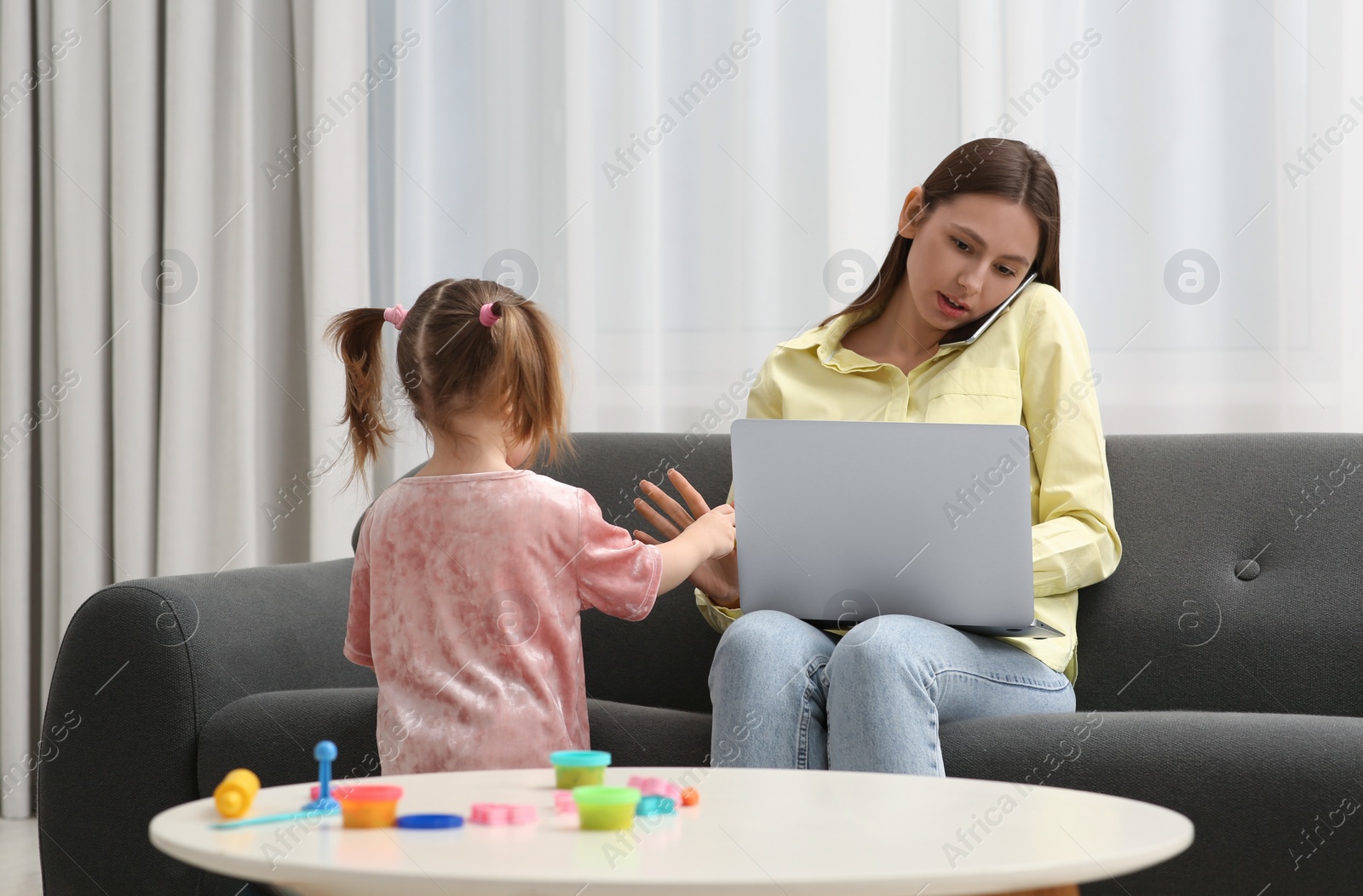 Photo of Little girl bothering her mother at home. Woman working remotely in living room