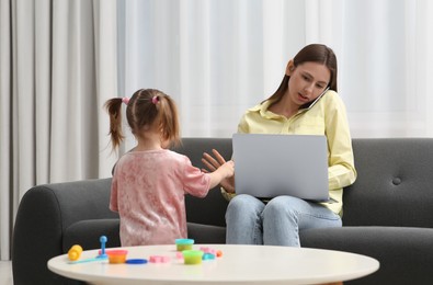 Photo of Little girl bothering her mother at home. Woman working remotely in living room