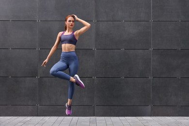 Photo of Beautiful woman in gym clothes jumping near dark grey wall on street, space for text
