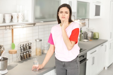 Photo of Young woman with sensitive teeth and glass of cold water in kitchen