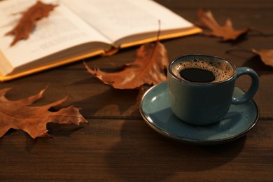 Cup of aromatic coffee, book and autumn leaves on wooden table. Space for text