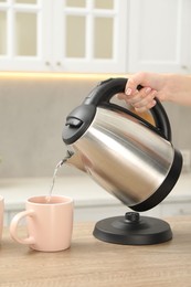 Photo of Woman pouring hot water from electric kettle into cup in kitchen, closeup