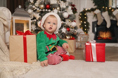 Cute baby wearing Santa's elf clothes near Christmas tree at home