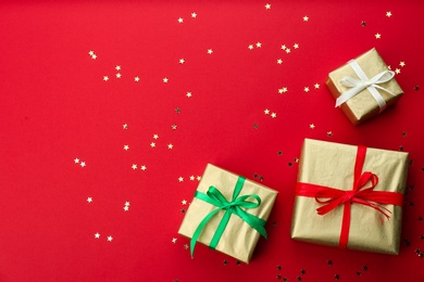 Photo of Christmas gift boxes and shiny confetti on red background, flat lay. Space for text