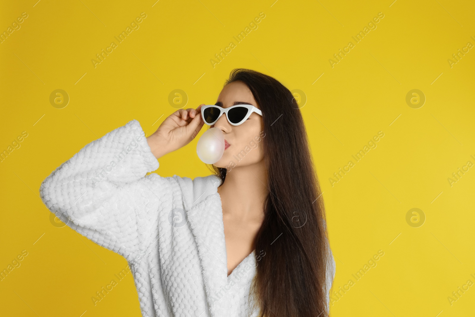 Photo of Young woman in bathrobe and sunglasses blowing chewing gum on yellow background