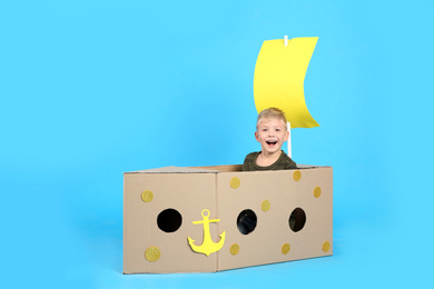 Photo of Little child playing with ship made of cardboard box on light blue background