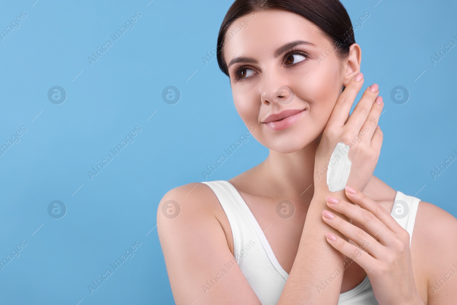 Photo of Beautiful woman with smear of body cream on her hand against light blue background, space for text