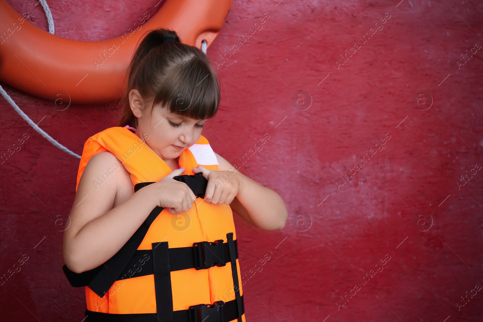 Photo of Little girl putting on orange life vest near red wall with safety ring. Space for text