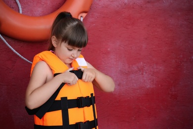 Photo of Little girl putting on orange life vest near red wall with safety ring. Space for text