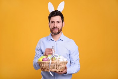Portrait of happy man in cute bunny ears headband holding wicker basket with Easter eggs on orange background