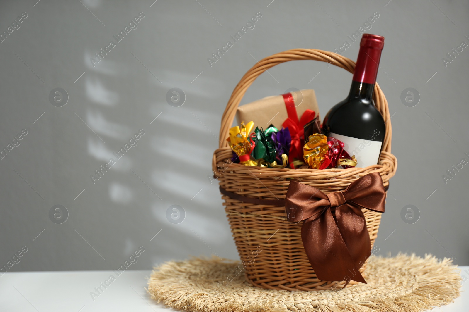 Photo of Wicker gift basket with food and wine on white table against grey background. Space for text