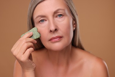 Woman massaging her face with jade gua sha tool on brown background
