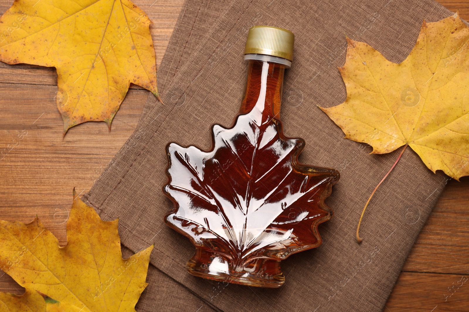 Photo of Bottle of tasty maple syrup and dry leaves on wooden table, flat lay