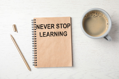 Notebook with phrase NEVER STOP LEARNING, pen and cup of coffee on white wooden background, flat lay