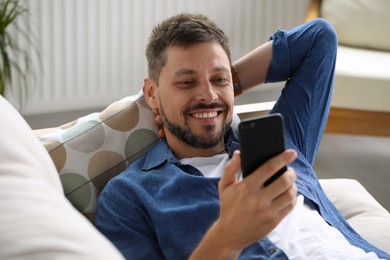 Photo of Happy man laying on sofa and using smartphone at home