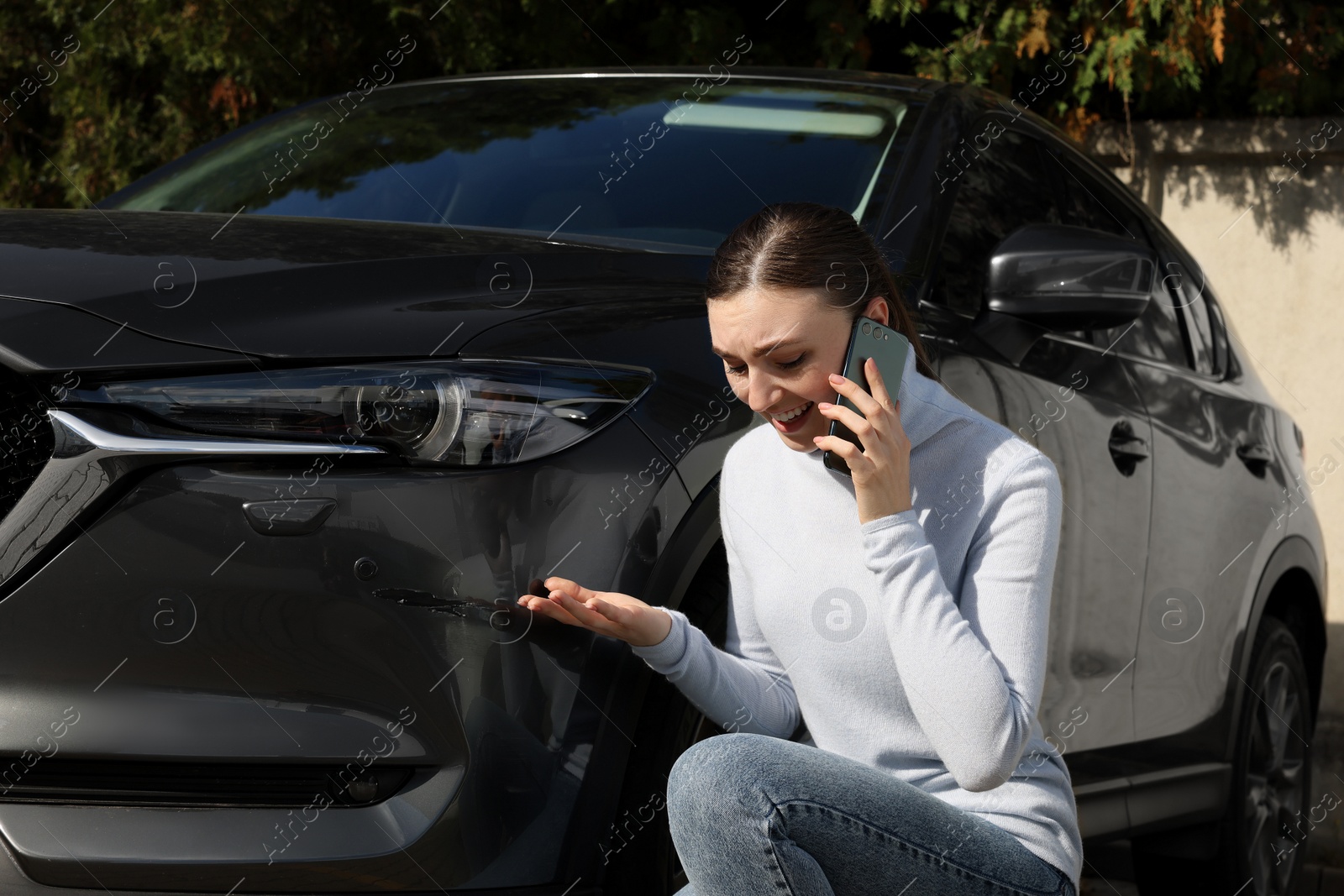 Photo of Stressed woman talking on phone near car with scratch outdoors