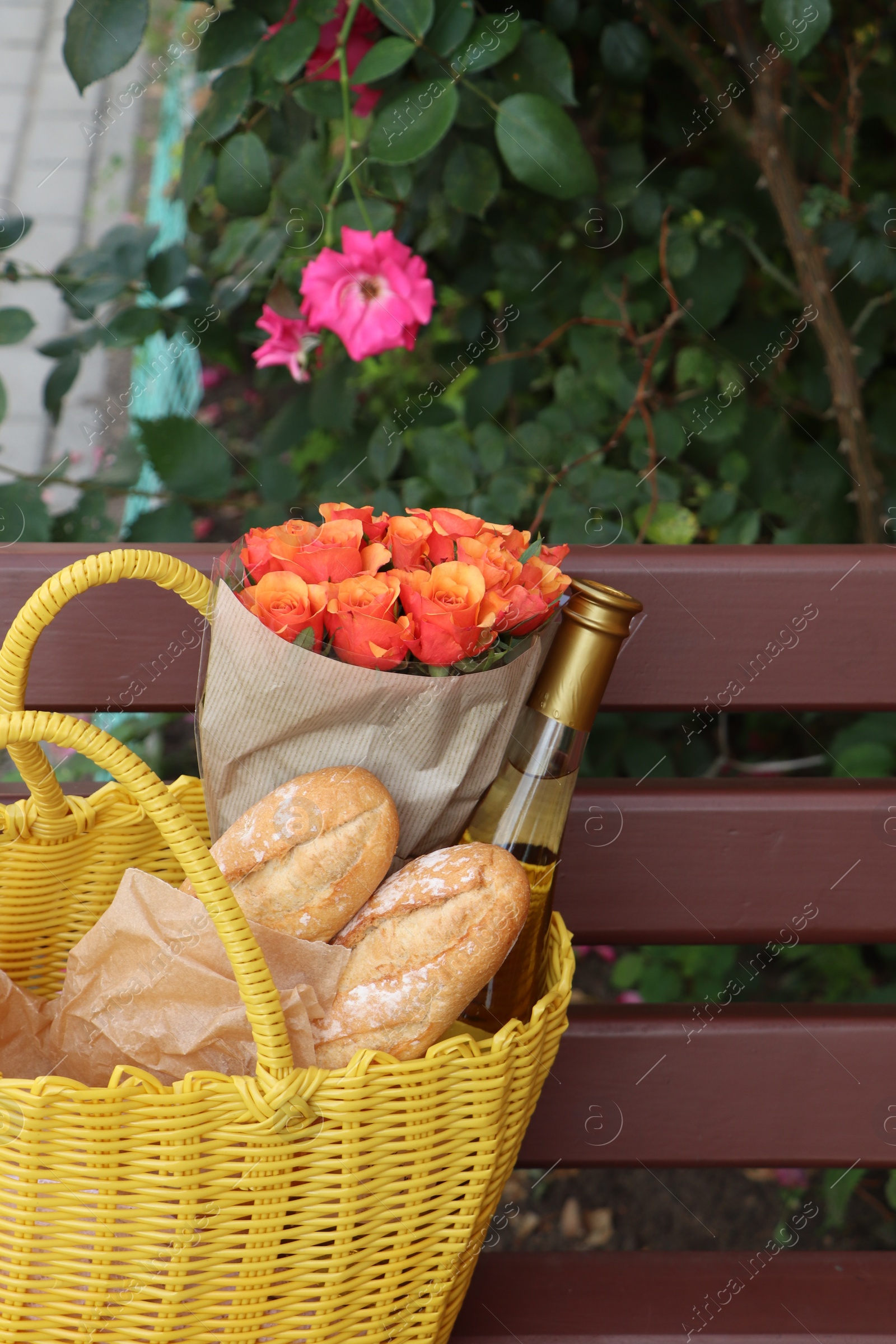 Photo of Beautiful roses, bottle of wine and baguettes in yellow wicker bag on bench outdoors