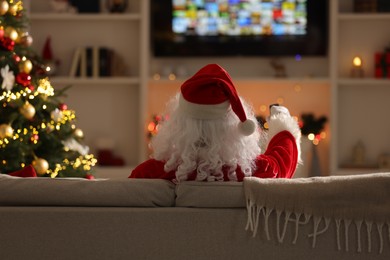 Photo of Merry Christmas. Santa Claus with remote control watching TV on sofa at home, back view