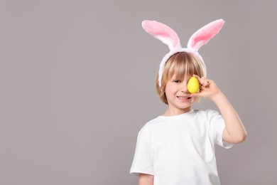 Happy boy in bunny ears headband holding painted Easter egg on grey background. Space for text