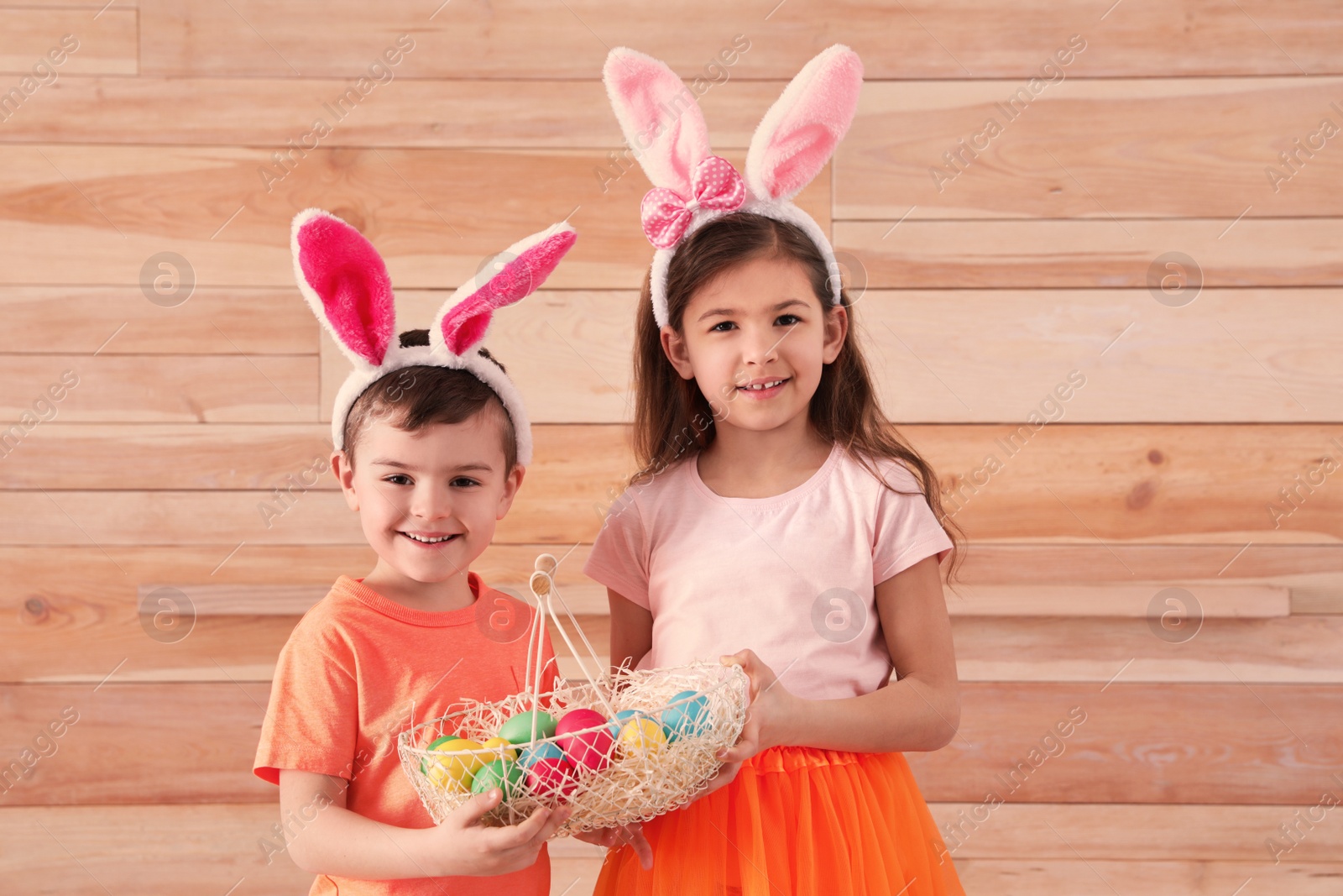 Photo of Cute children in bunny ears headbands holding basket with Easter eggs against wooden background