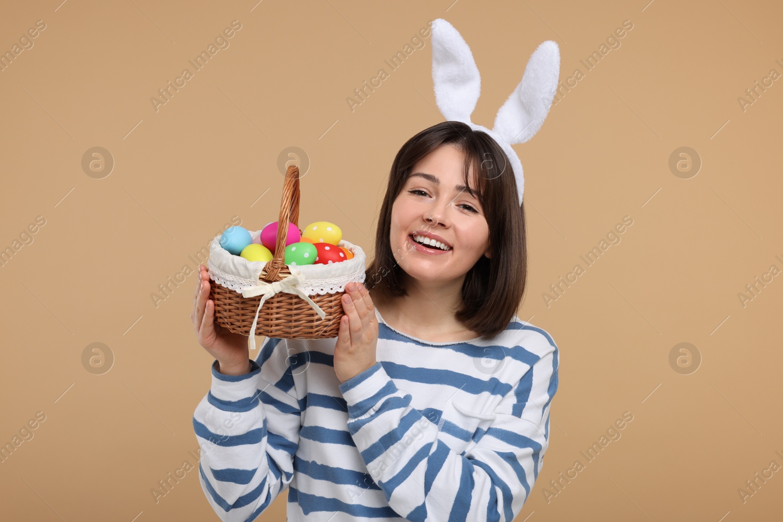 Photo of Easter celebration. Happy woman with bunny ears and wicker basket full of painted eggs on beige background