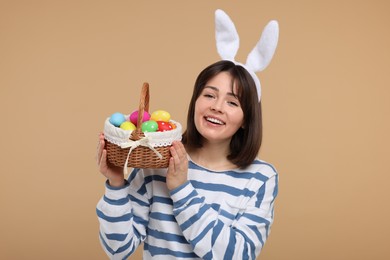 Easter celebration. Happy woman with bunny ears and wicker basket full of painted eggs on beige background