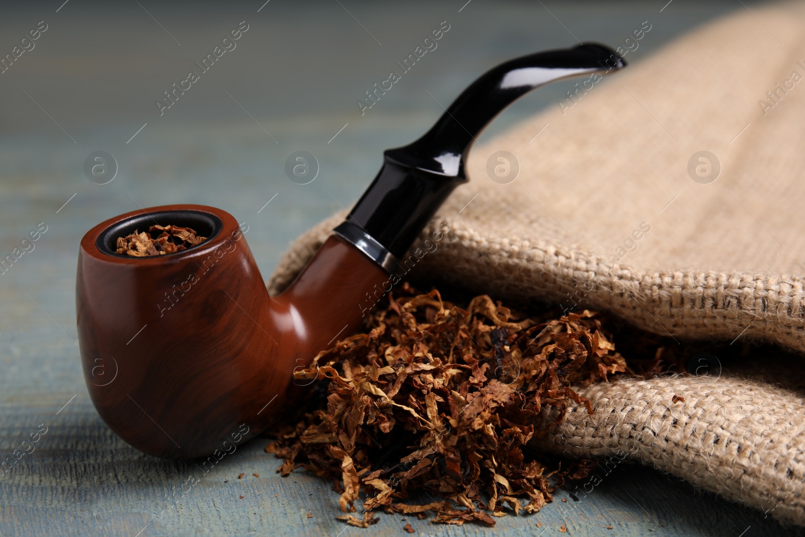 Photo of Smoking pipe, dry tobacco and sackcloth on blue wooden table, closeup