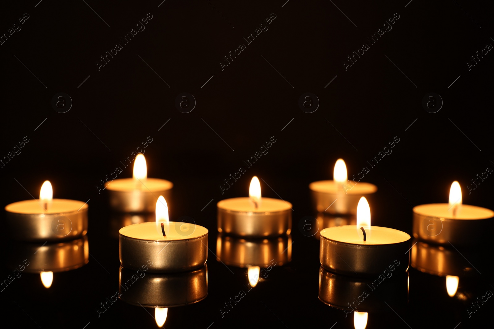 Photo of Burning candles on mirror surface in darkness, closeup