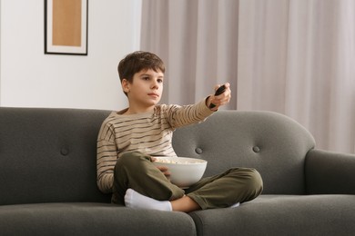 Photo of Little boy holding bowl of popcorn and changing TV channels with remote control on sofa at home