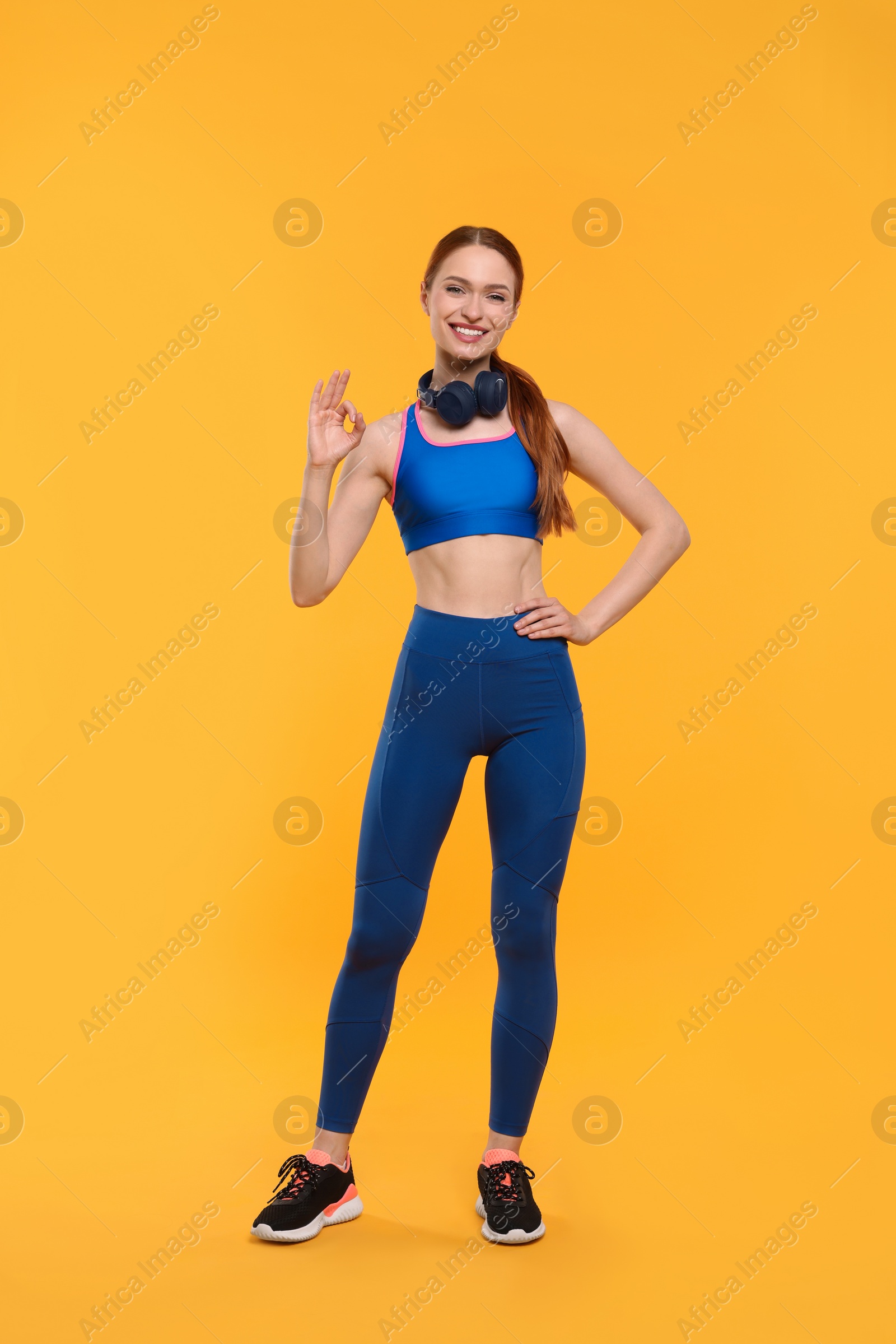 Photo of Young woman in sportswear and headphones showing ok gesture on yellow background