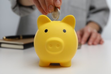 Photo of Woman putting coin into yellow piggy bank at white table, closeup