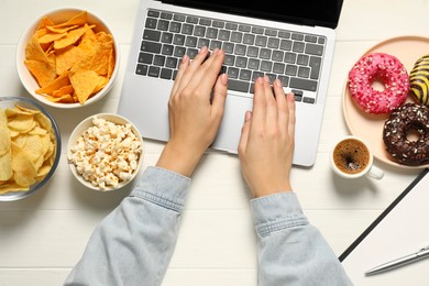 Photo of Bad eating habits. Woman using laptop surrounded by different snacks at white wooden table, top view