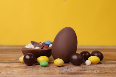 Photo of Delicious chocolate eggs and colorful candies on wooden table against yellow background