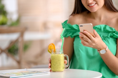 Young woman using mobile phone while drinking tasty healthy smoothie at table, indoors