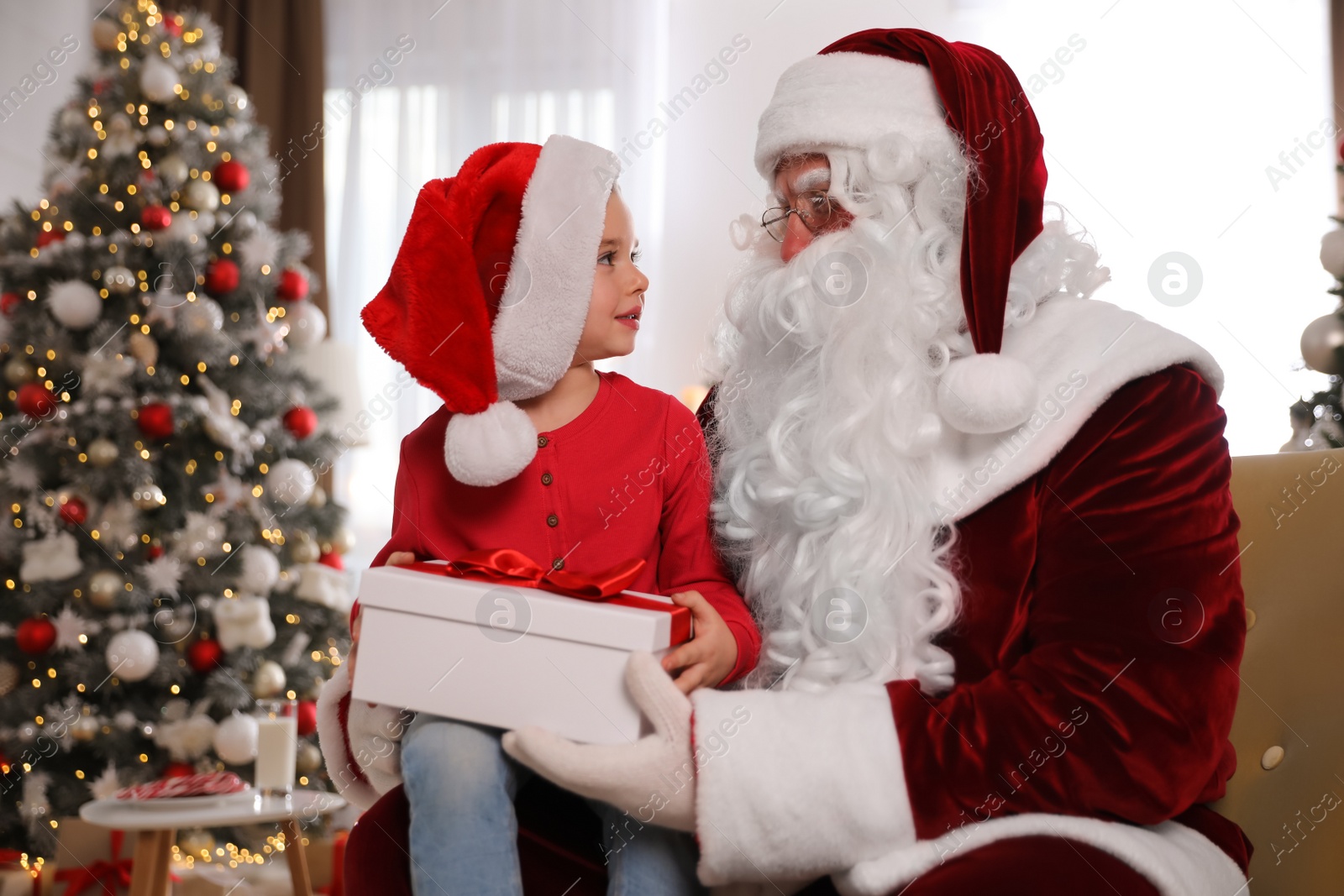 Photo of Santa Claus giving present to little girl in room decorated for Christmas