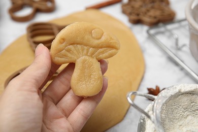 Photo of Woman holding unbaked mushroom shaped cookie at white table, closeup