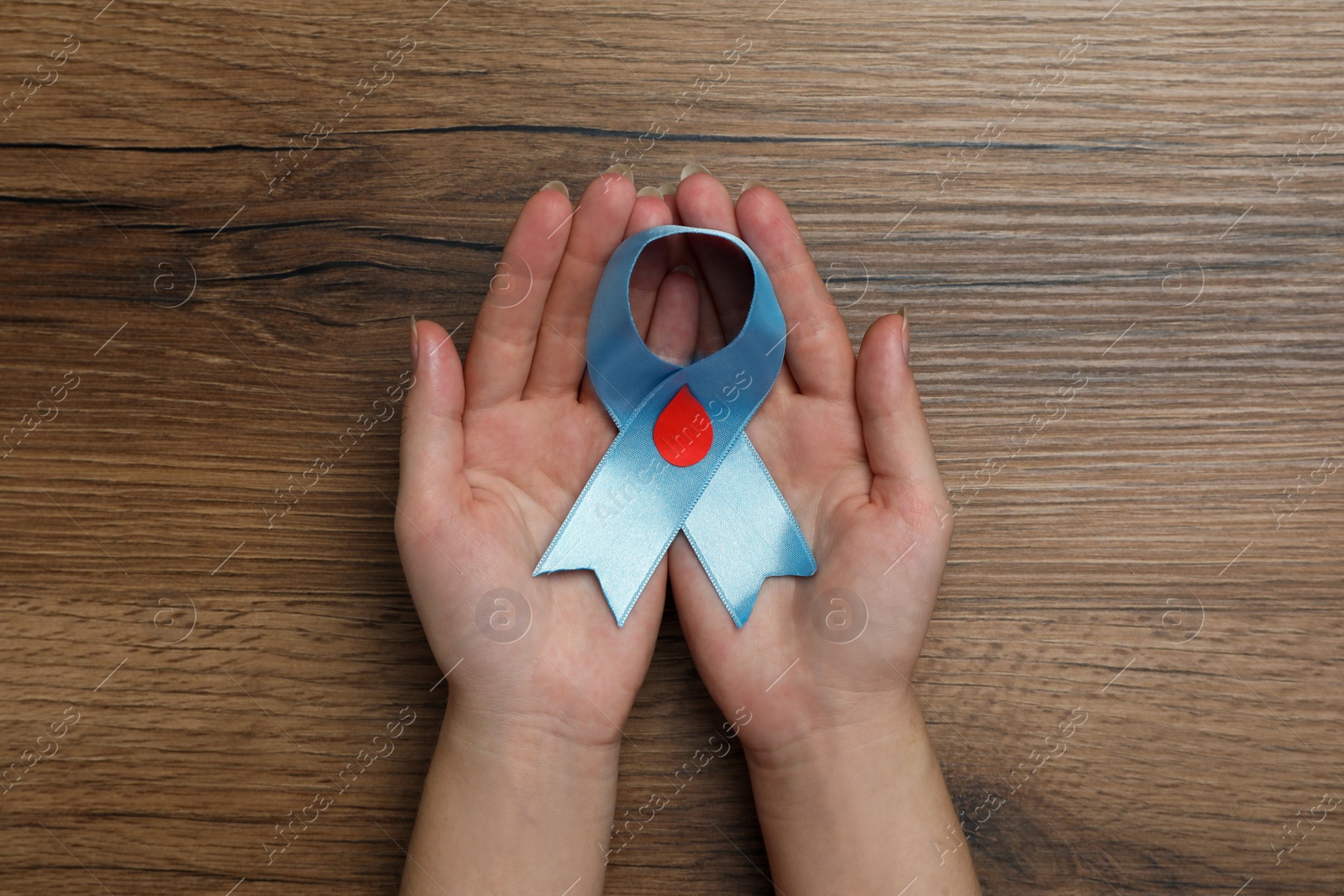 Photo of Woman holding light blue ribbon with paper blood drop at wooden table, top view. Diabetes awareness