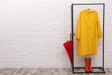 Photo of Stylish rack with umbrella, raincoat and rubber boots near white brick wall. Space for text