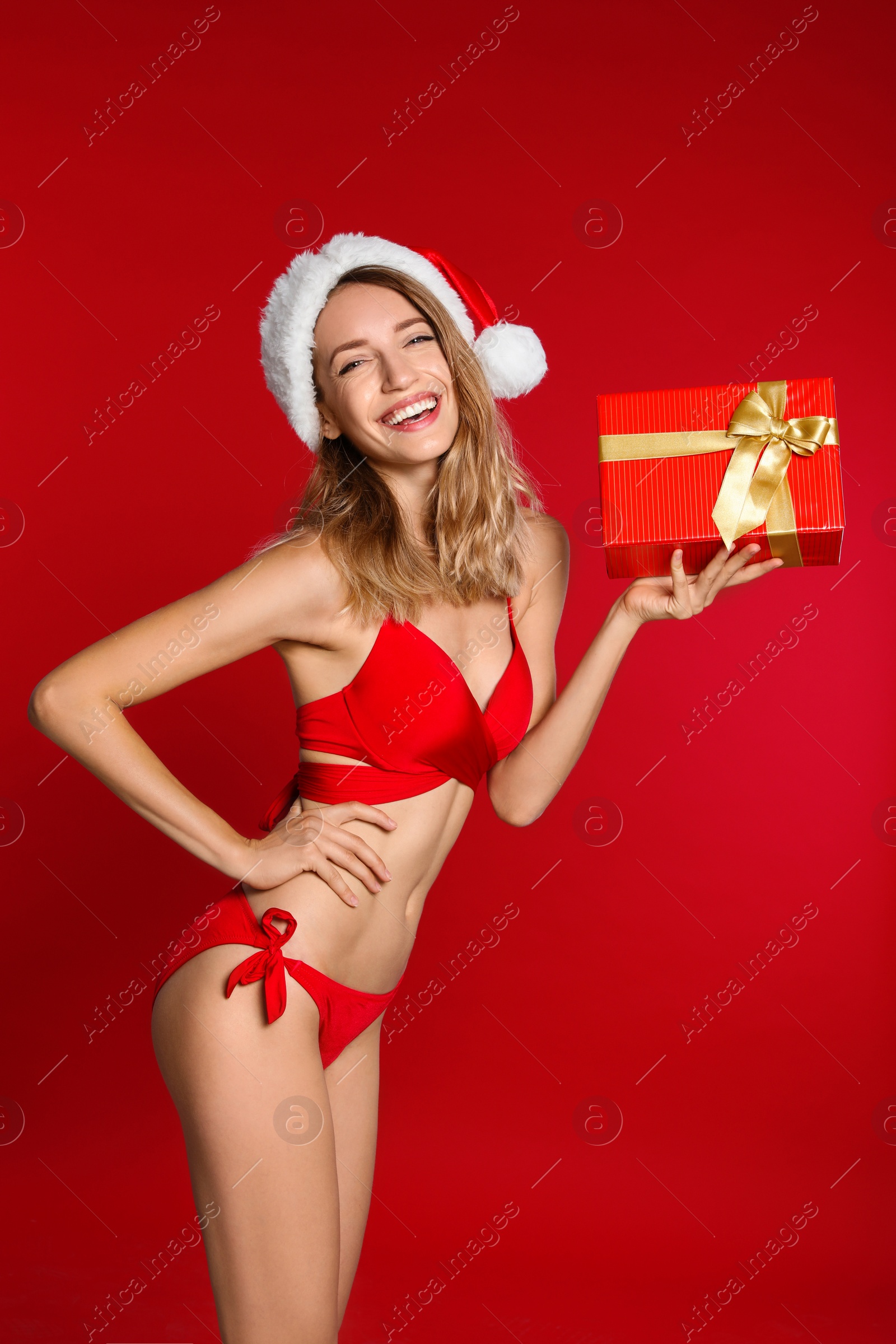 Photo of Happy young woman in bikini and Santa hat with gift box on red background. Christmas celebration