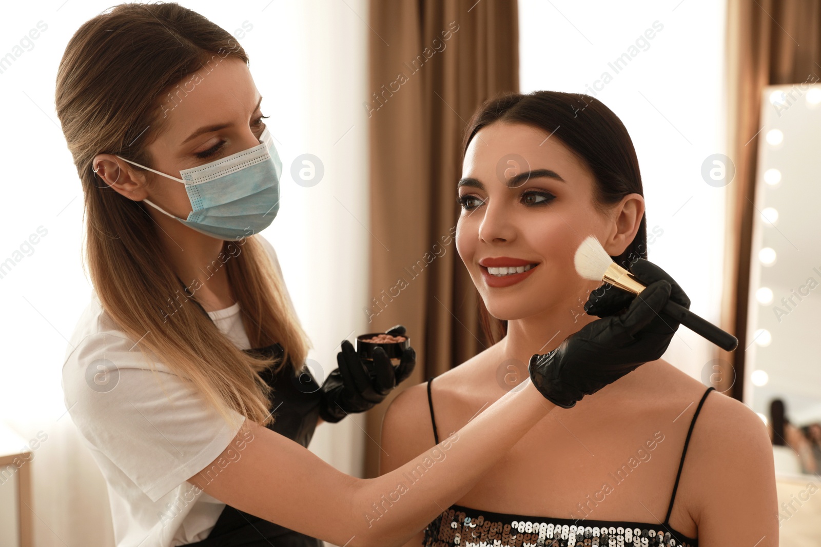 Photo of Makeup artist in protective mask and gloves working with woman indoors. Preventive measures during COVID-19 pandemic