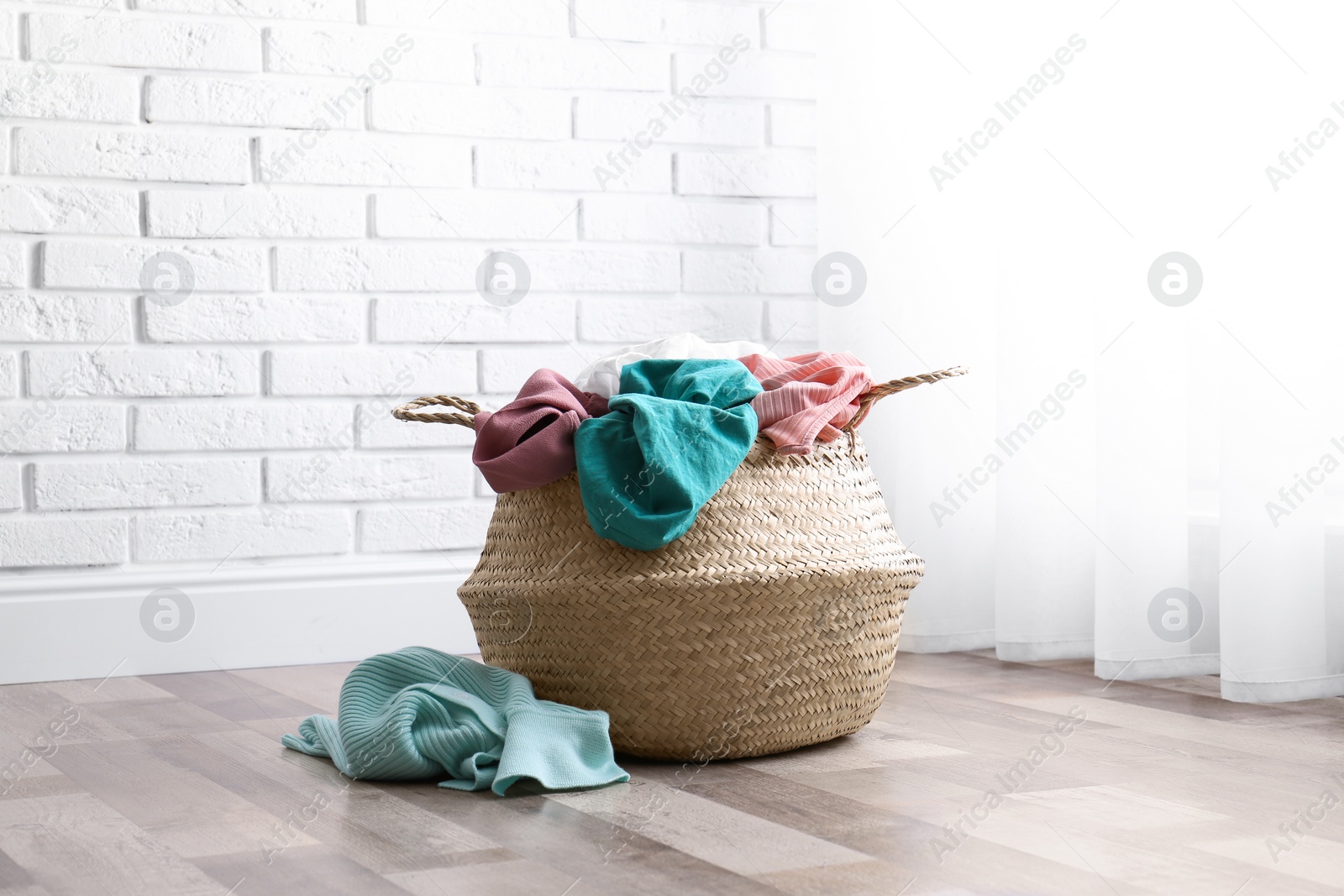 Photo of Wicker basket with dirty laundry on floor near window