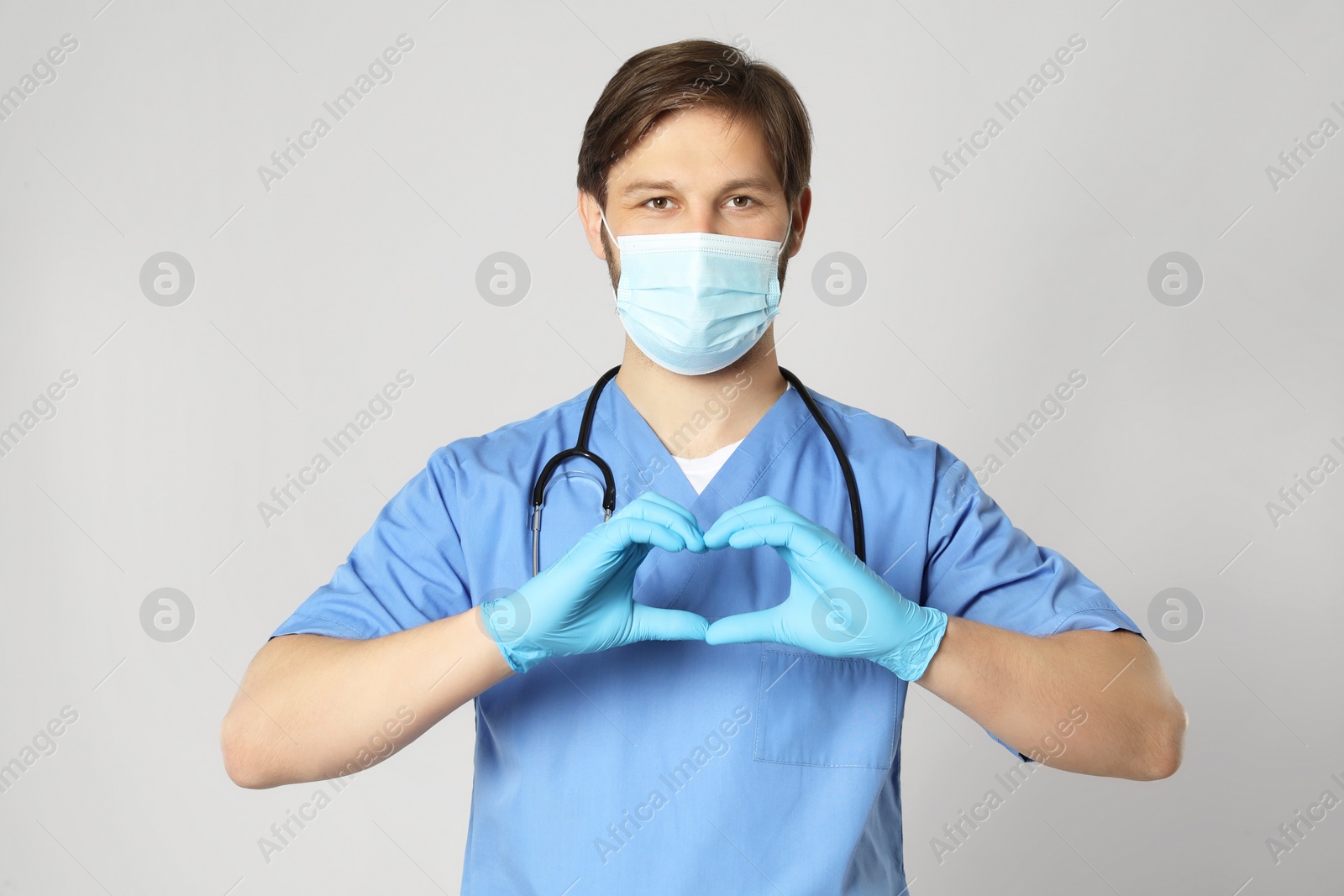 Photo of Doctor or medical assistant (male nurse) with protective mask and stethoscope making heart on light grey background