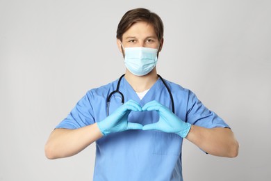 Photo of Doctor or medical assistant (male nurse) with protective mask and stethoscope making heart on light grey background