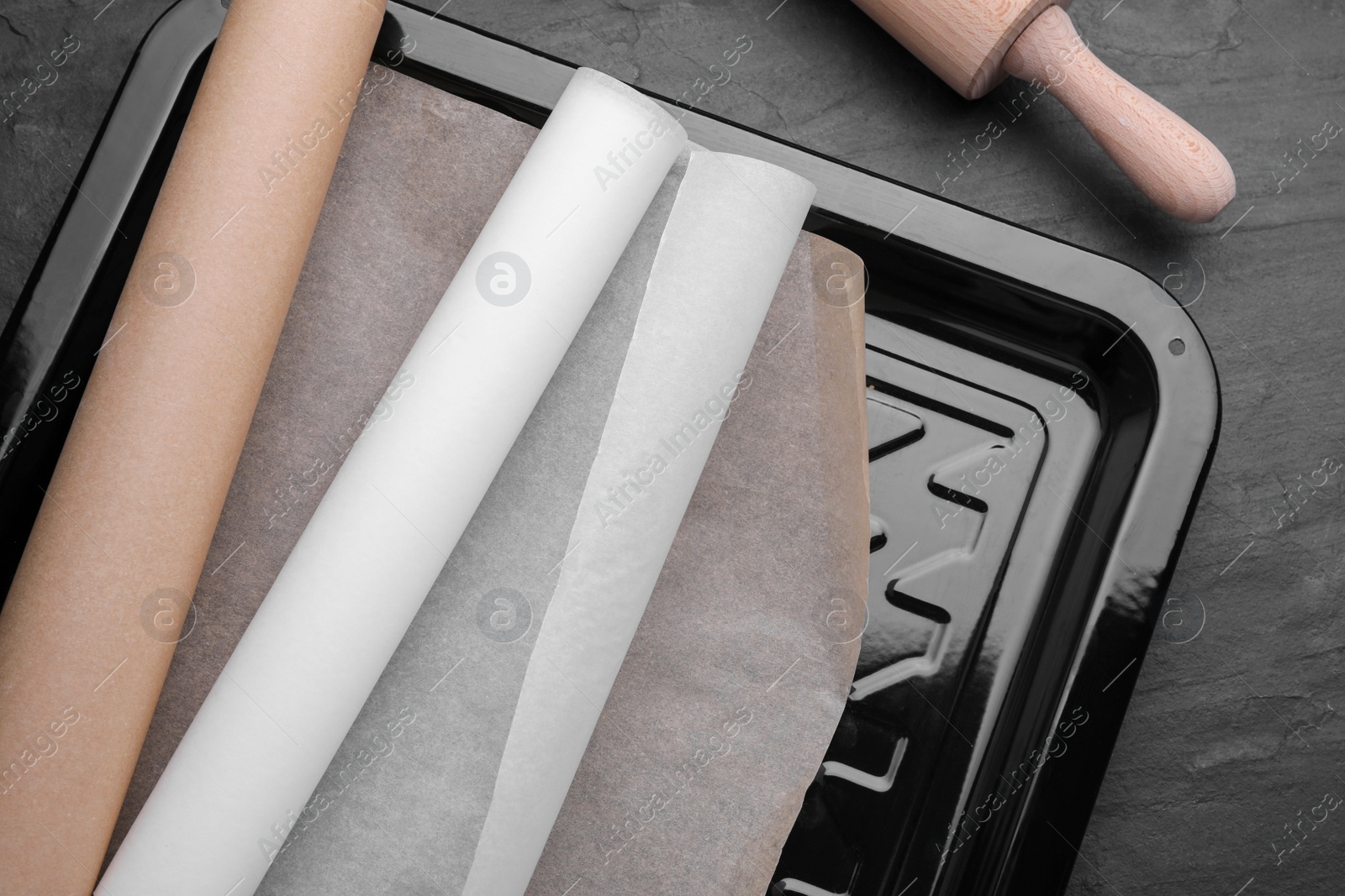 Photo of Baking pan with rolls of parchment paper on black table, top view