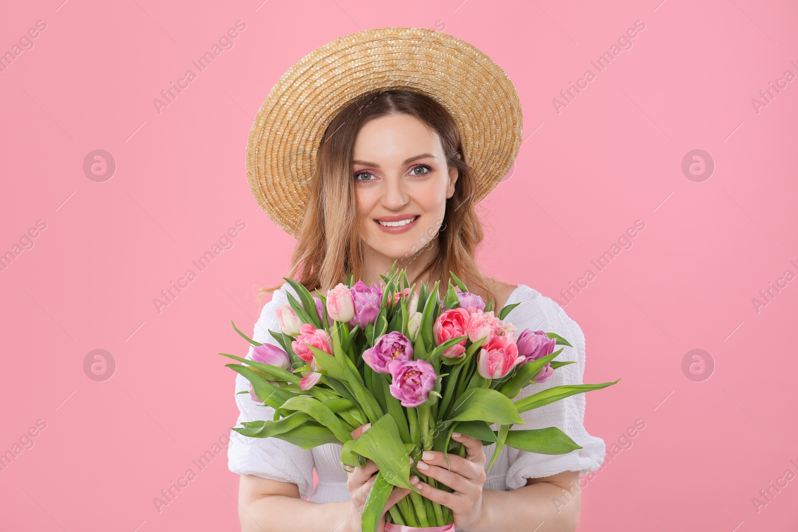 Photo of Happy young woman in straw hat holding bouquet of beautiful tulips on pink background