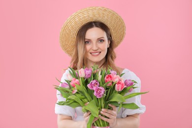 Happy young woman in straw hat holding bouquet of beautiful tulips on pink background