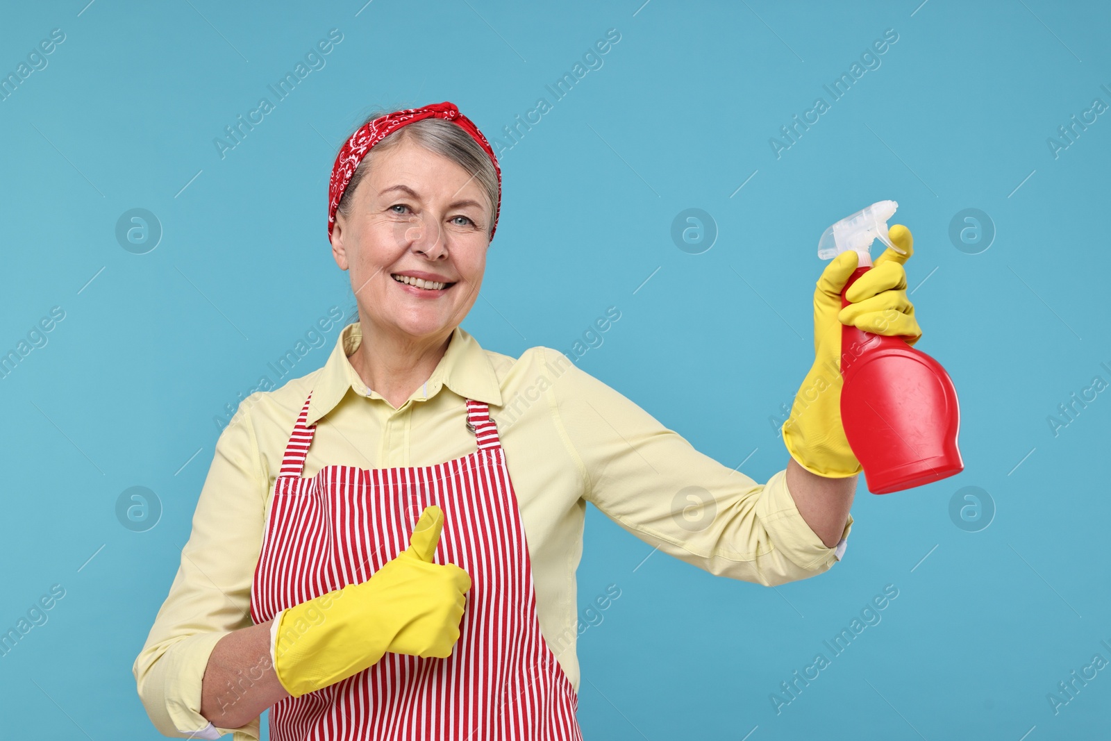 Photo of Happy housewife with spray bottle showing thumbs up on light blue background