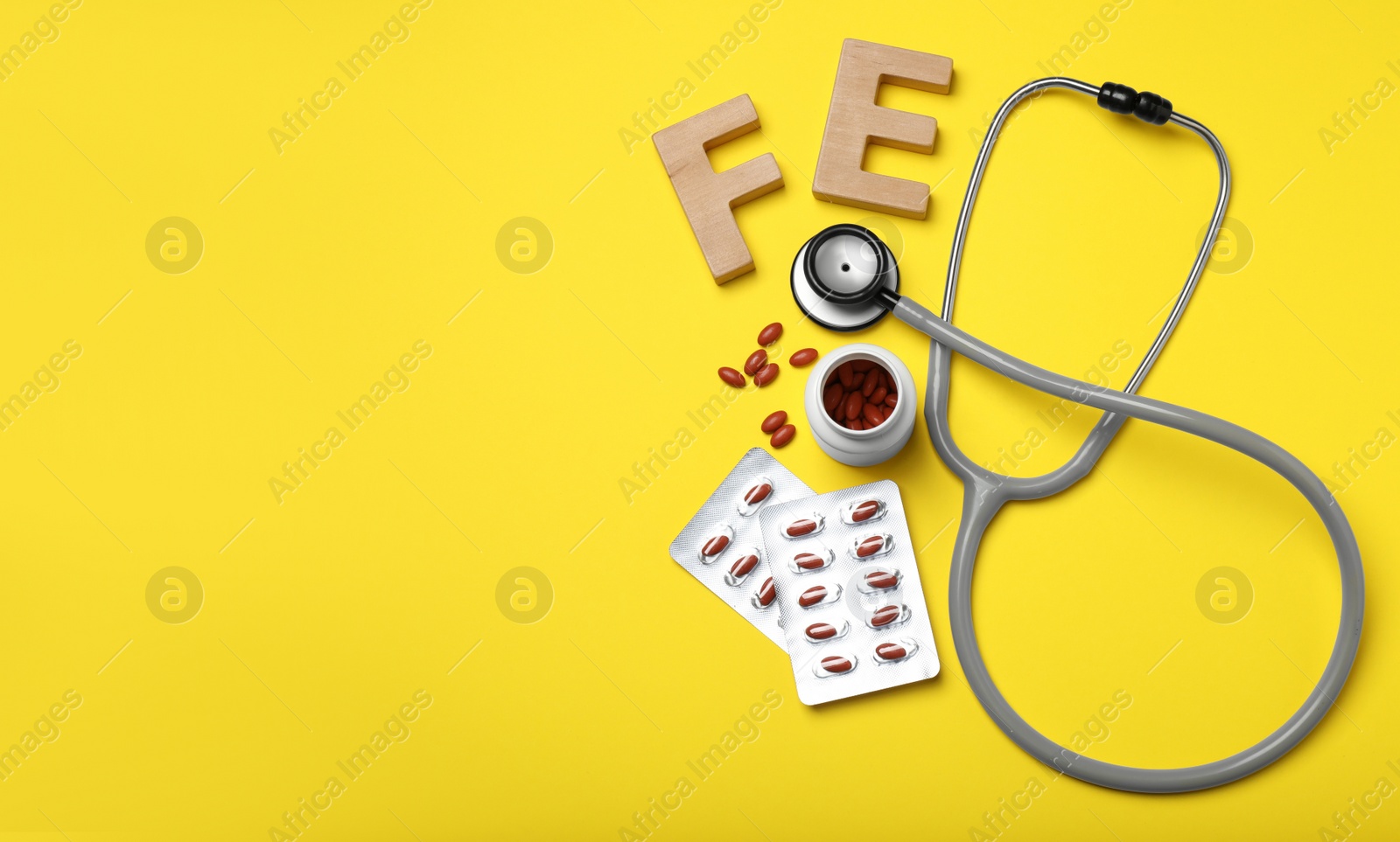 Photo of Wooden letters FE, pills and stethoscope on yellow background, flat lay with space for text. Anemia treatment